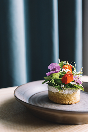 Closeup of a vegan crumpet dish with beautiful florals and green leaves. The plate sits on a wooden table with blue curtain in the background.