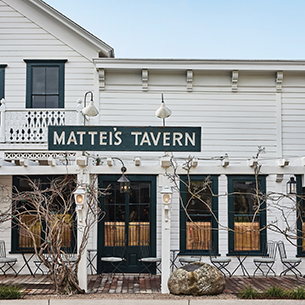 White store front of Mattei’s Tavern restaurant. There are several windows on the wooden building as well as tables and chairs on the sidewalk in front of the building. 