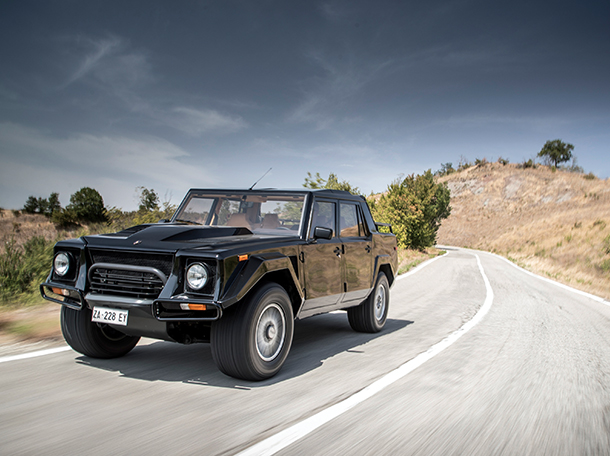 A black Lamborghini LM002 1986 SUV speeds down a deserted country road lined with tall trees.