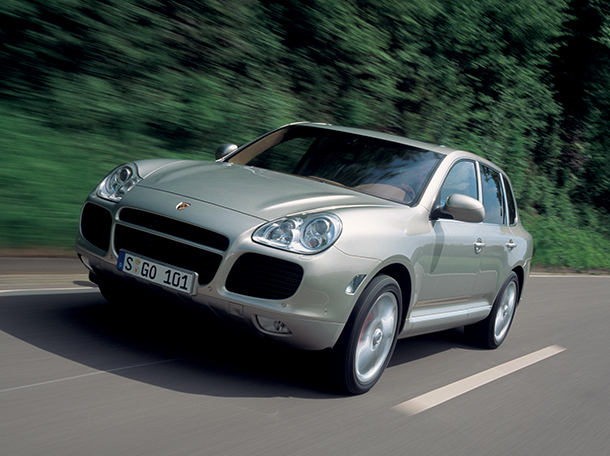A silver Porsche Cayenne 2002 SUV speeds down a winding road, with lush greenery in the background