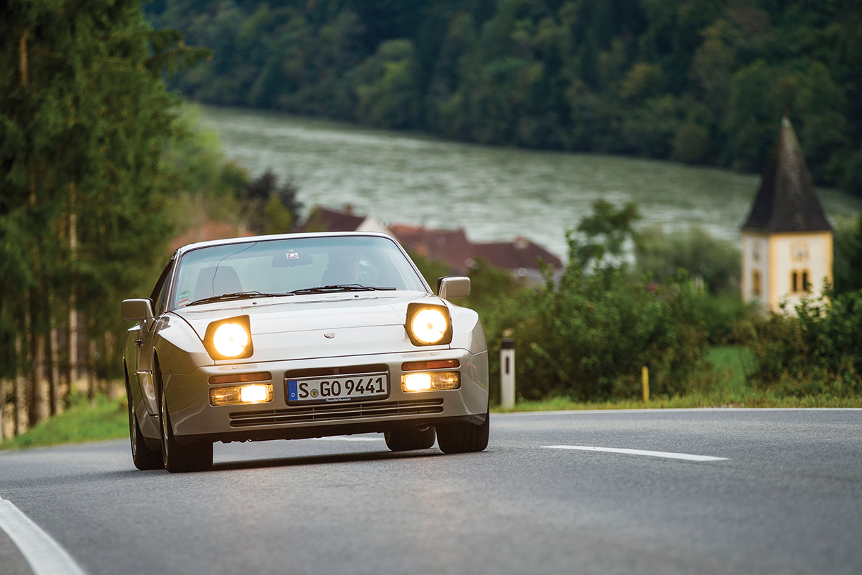 A classic silver Porsche 944 Turbo S speeds down a winding country road, surrounded by lush greenery.