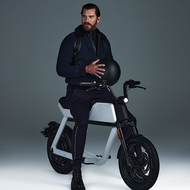 Man sitting on white Pave BK e-motorbike wearing dark clothes, boots and a black backpack while holding a black riding helmet.