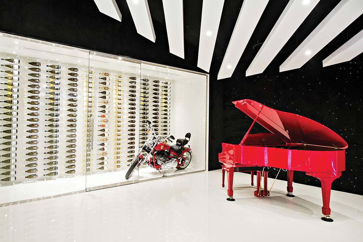A custom wine cellar with glass doors and a Ferrari motorcycle parked inside the case as well as a red grand piano outside in the showroom.