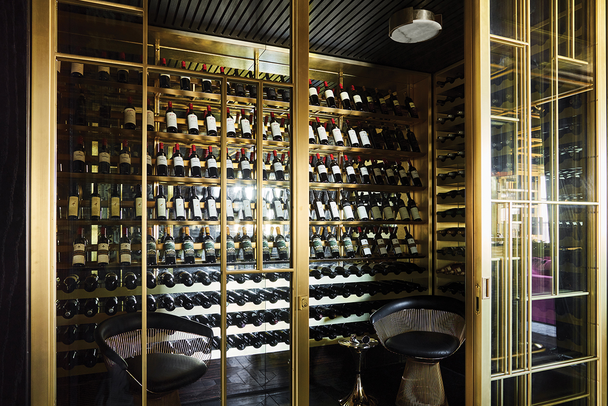A well-stocked, elegant wine cellar with brass-trimmed sliding glass doors, custom brass wine shelving, designer Art Deco style chairs and chandeliers.