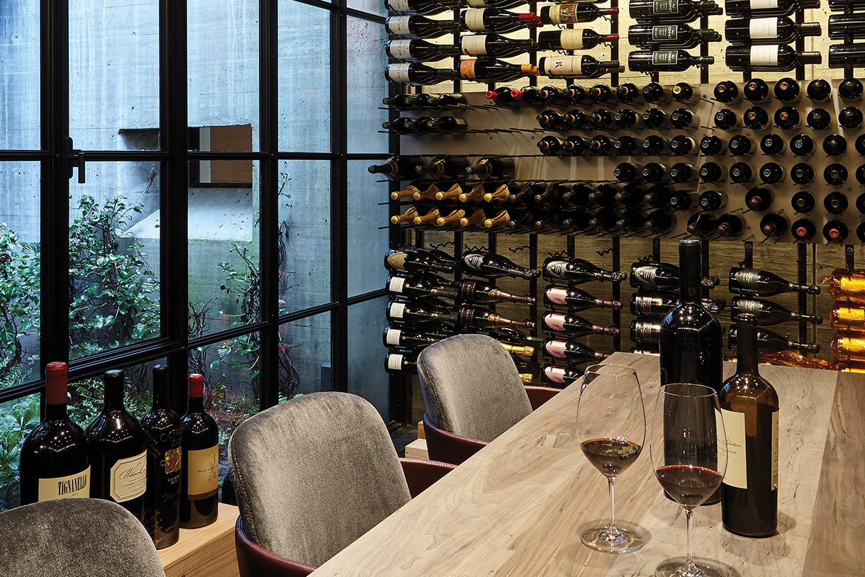 A sophisticated and contemporary wine cellar with warm wood and concrete accents display a collection of wines against the wall. A large wooden table, floor-to-ceiling glass wall overlooking a courtyard, and slatted wood ceiling provide a luxurious, yet natural aesthetic.