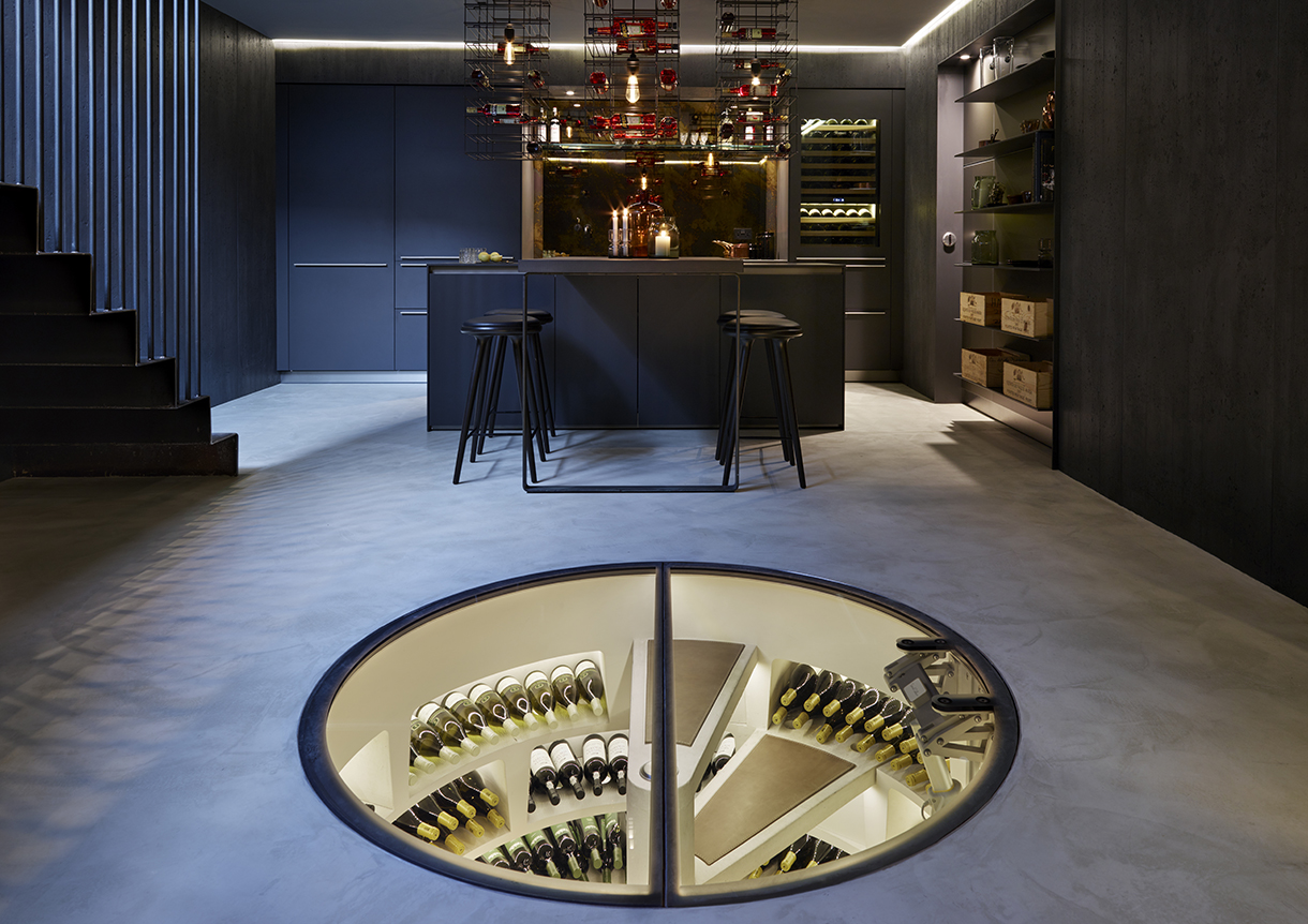 A modern kitchen with dark sleek cabinets and appliances. A circular, glass-walled wine cellar built into the floor with a spiral staircase.
