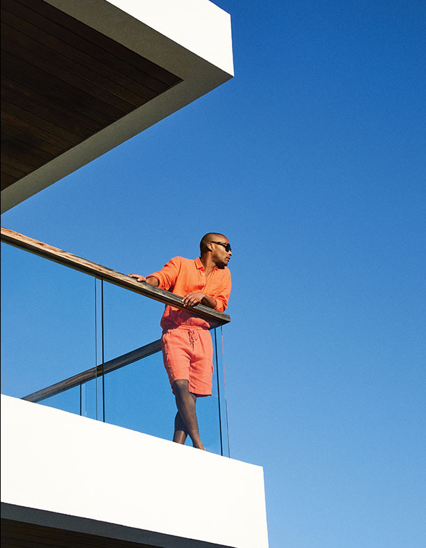 Man standing on balcony overlooking view wearing orange linen shirt and shorts
