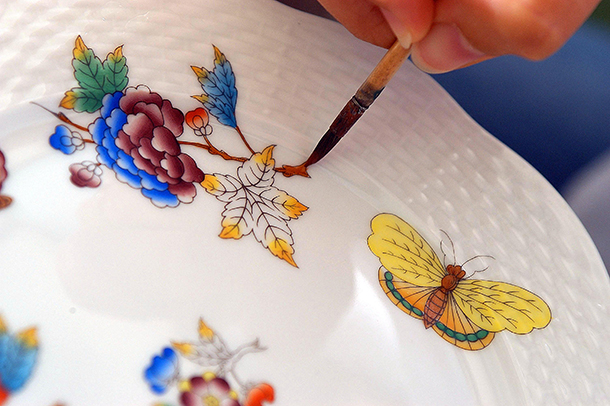 Person hand-painting a vibrant and exquisite floral motif of flowers, leaves and butterflies on a white porcelain plate.