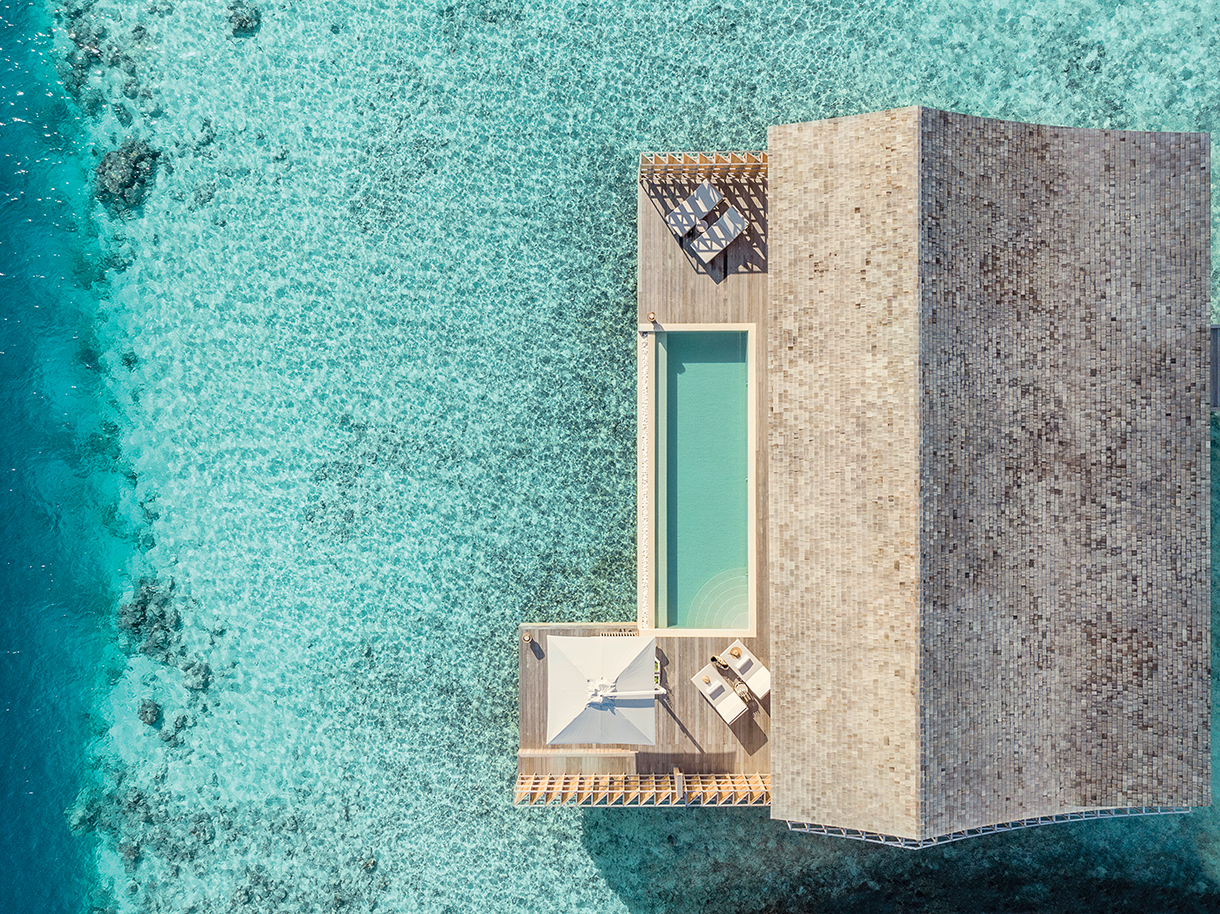 Beautiful aerial view of an over-water residence at the Kudadoo Maldives Private Island with an outdoor deck and infinity plunge pool surrounded by crystal-clear turquoise water.