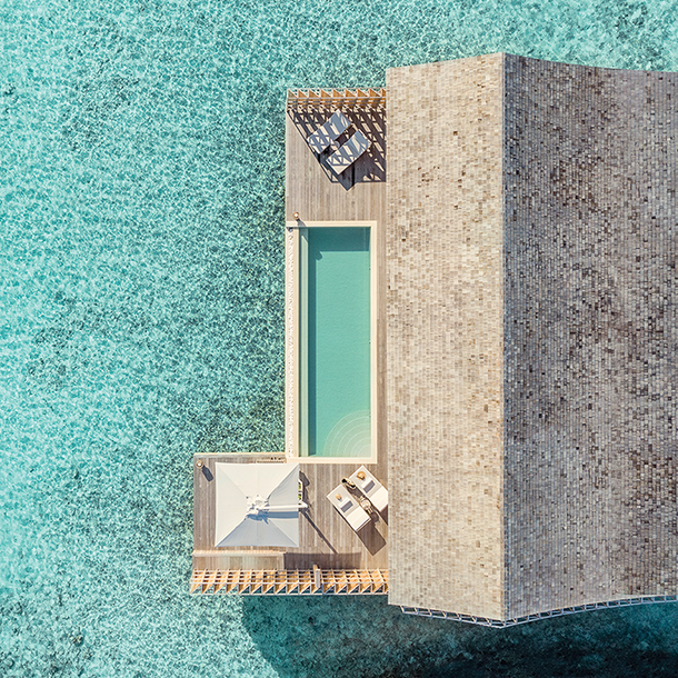 Beautiful aerial view of an over-water residence at the Kudadoo Maldives Private Island with an outdoor deck and infinity plunge pool surrounded by crystal-clear turquoise water.