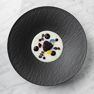 Beautifully plated pudding with dark berries and colorful flowers served on black textured dish