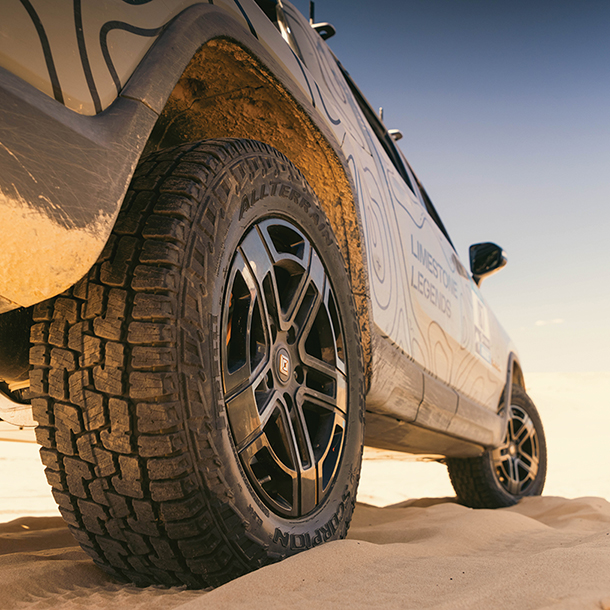 Closeup of the rear wheel, tire and side of gray Rivian R1T rally truck parked on sand