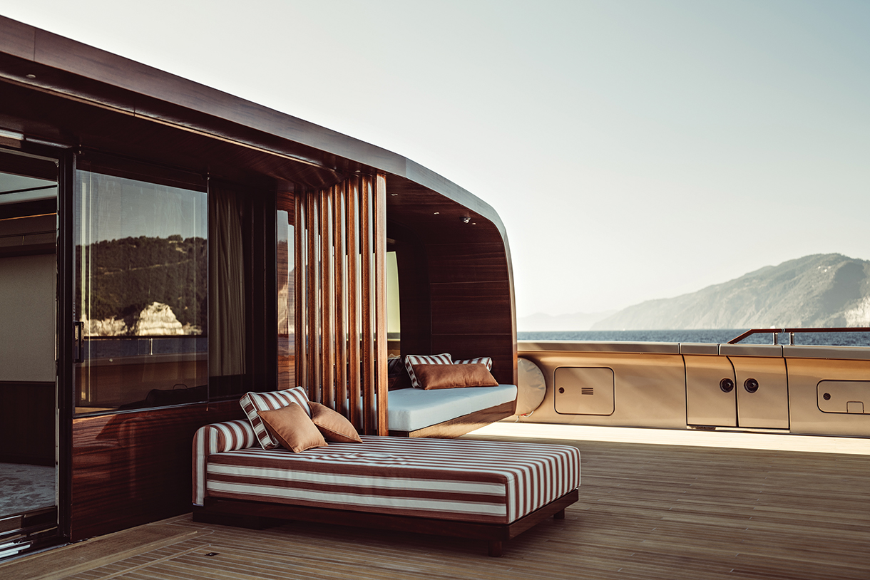 The spacious wooden deck of a luxury catamaran super yacht has cabins with floor-to-ceiling windows and chaise lounges with pillows. The sea and mountains are visible in the background.