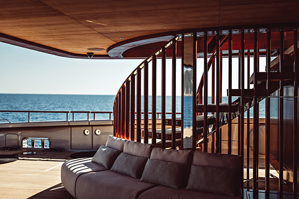 A luxury catamaran super yacht with a spiral staircase made of wood and metal, and a curved sofa with pillows. The sea is visible in the background. The yacht is designed by Axel de Beaufort and Guillaume Verdier for Perini Navi. 