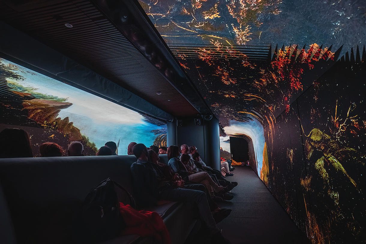 The interior of a boat that has been converted into a museum. It shows the main exhibition space, which is a long, narrow room with a high ceiling. The walls are covered in projections of paintings and other works of art. There are people sitting on benches in the room, watching the projections.