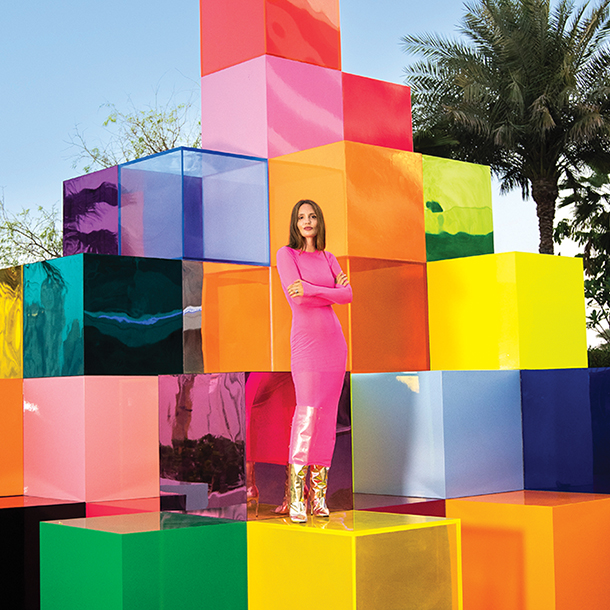 Woman wearing pink outfit standing on top of a large yellow cube among a stack of other colorful cubes which form an abstract art piece