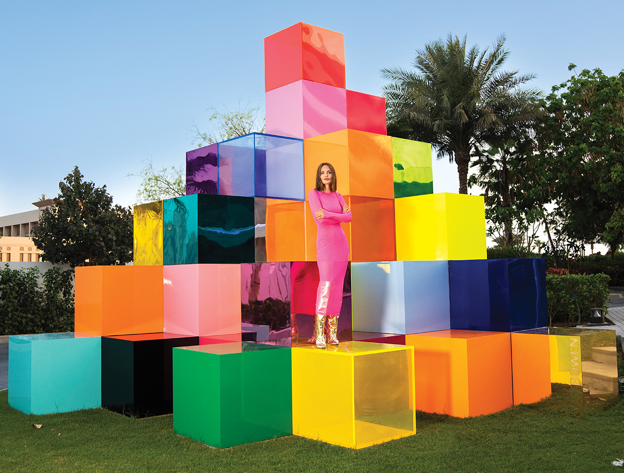 Woman wearing pink outfit standing on top of a large yellow cube among a stack of other colorful cubes which form an abstract art piece