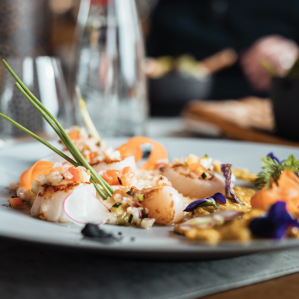 A plate of seared scallops with colorful vegetables and a garnish of chives. The scallops are arranged on a bed of pureed vegetables and are accompanied by small edible flowers.