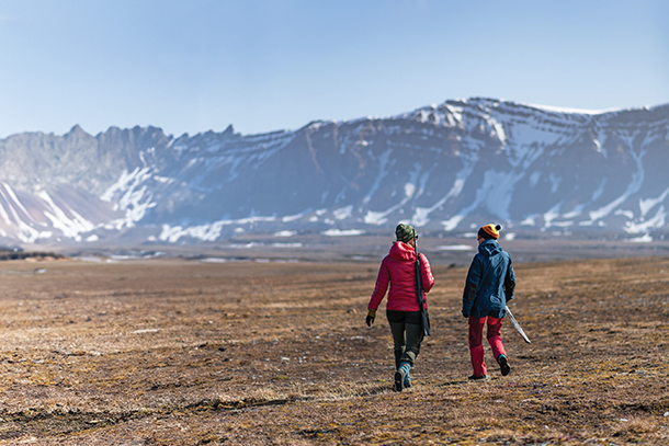 Two women wearing winter clothing walking in a field with snow covered mountains in the distance.