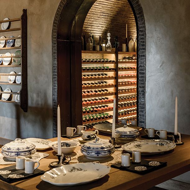 A dining table set for a meal with dishes and candles. In the background, a wine cellar is visible through an arched doorway. The wine cellar is filled with rows of wine bottles.