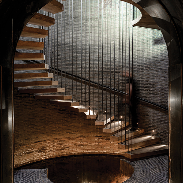 A circular staircase with wooden steps and metal railings. The staircase is surrounded by a brick wall and illuminated by soft lighting.