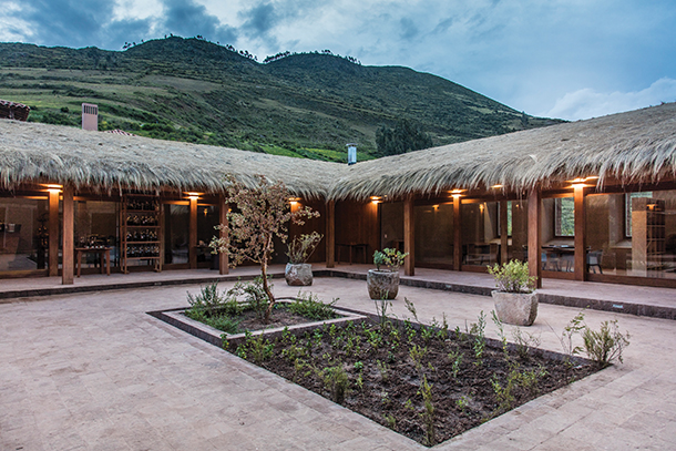 A traditional Peruvian building with a thatched roof and a central courtyard. The courtyard has a small garden and potted plants, with mountains visible in the background.