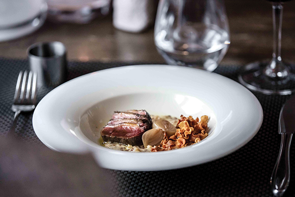 A plate of gourmet food with slices of reindeer meat and shallots. A glass of water is visible in the background.