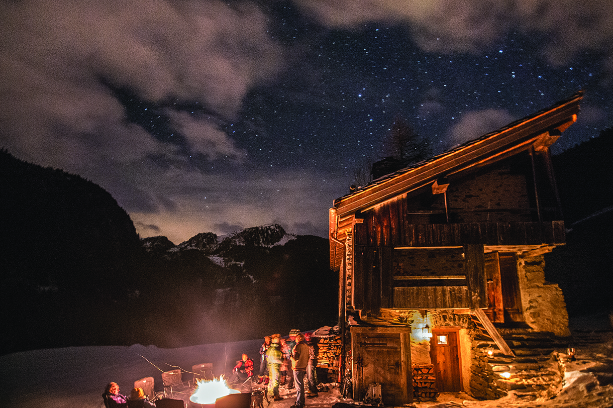  A wooden chalet with a warm fire outside. People are gathered around the fire, enjoying the night sky filled with stars. Mountains and clouds are visible in the background.