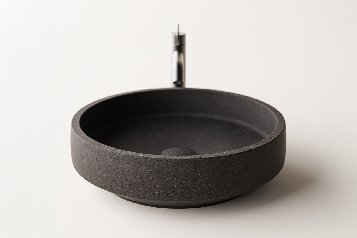 A round, dark gray stone countertop vessel used as a washbasin with a smooth, textured surface. An unique, modern chrome faucet is mounted on the countertop behind the vessel.