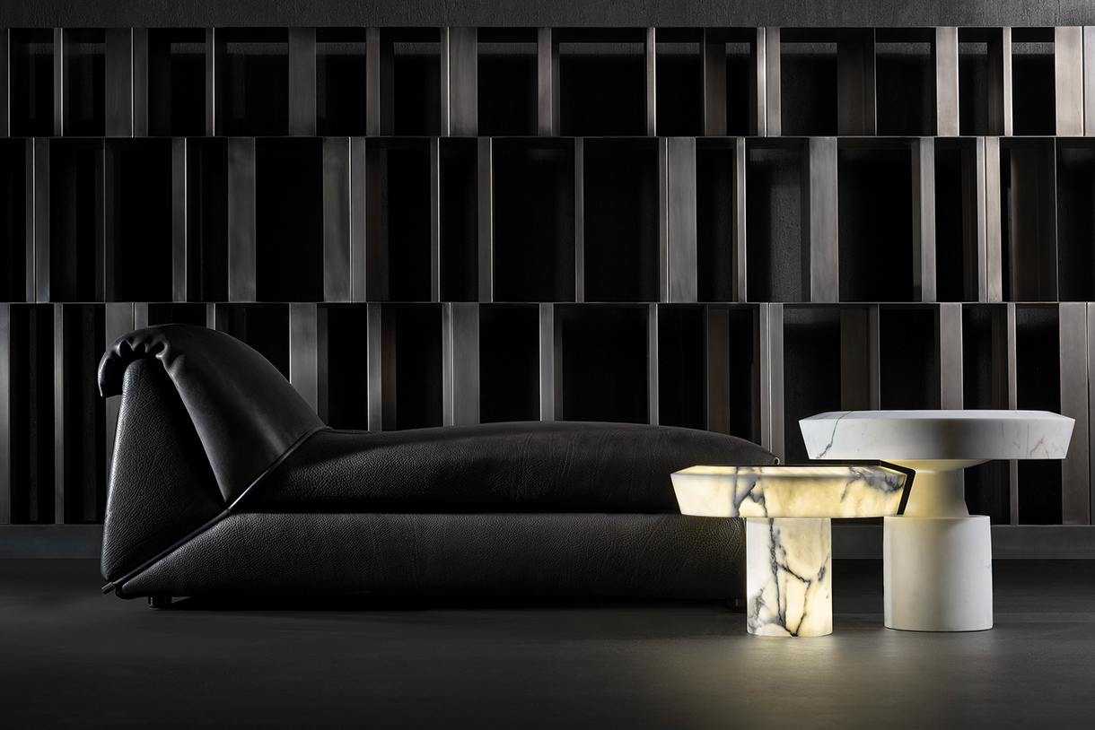 An illuminated marble side table with a unique, intersecting and geometric shape sits next to a chaise lounge. The lighting highlights the dramatic veining and structure of the stone. 