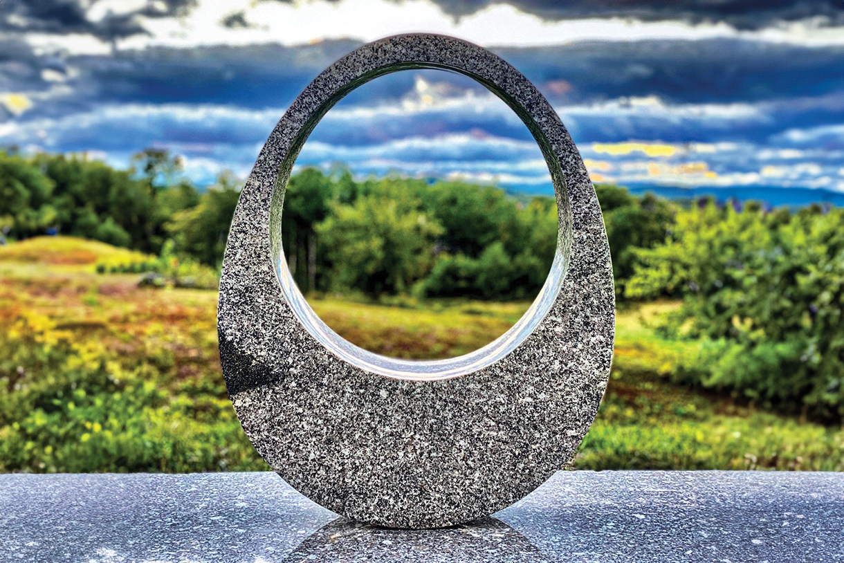 A large, oval granite sculpture with a hole in the center. The sculpture is placed on a stone platform with a scenic landscape of green fields and trees in the background.