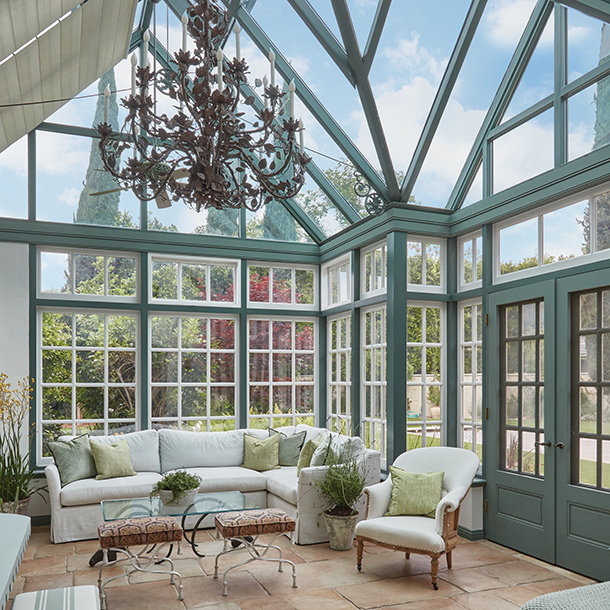 A sunroom with a glass ceiling and walls, featuring a large white sectional sofa, a coffee table, and armchairs. There is a chandelier hanging from the center of the room, and the windows have green frames.