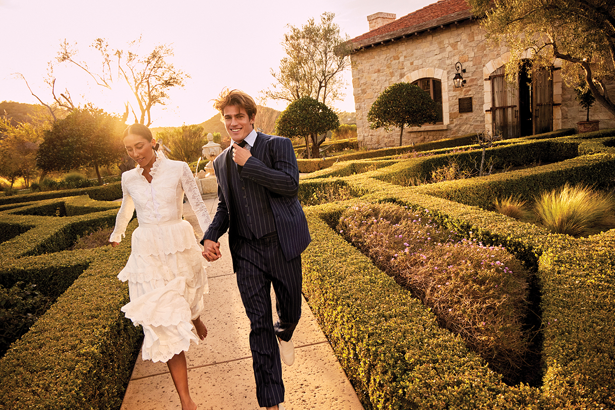 Woman dressed in a white dress holding hands and running with a man dressed in a three-piece, pin-striped navy suit in a beautiful French countryside-style garden 