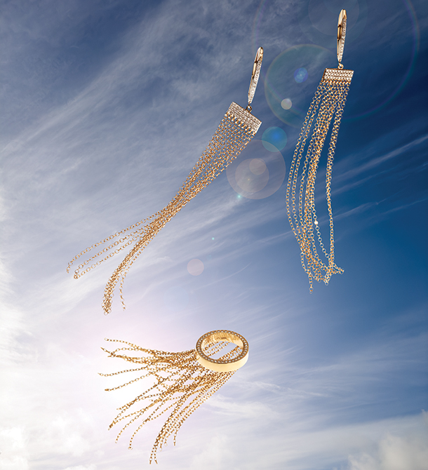  A pair of gold and diamond hoop earrings with a diamond bar and hanging chain design and a gold and diamond ring with hanging chains floating in a clear blue sky. 
