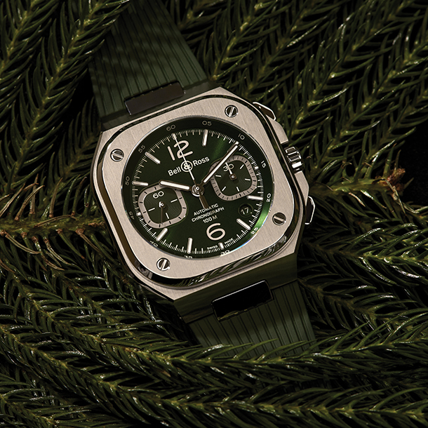 Steel watch with sporty square shaped case and dark green dial and rubber strap on a pile of green pine needles.