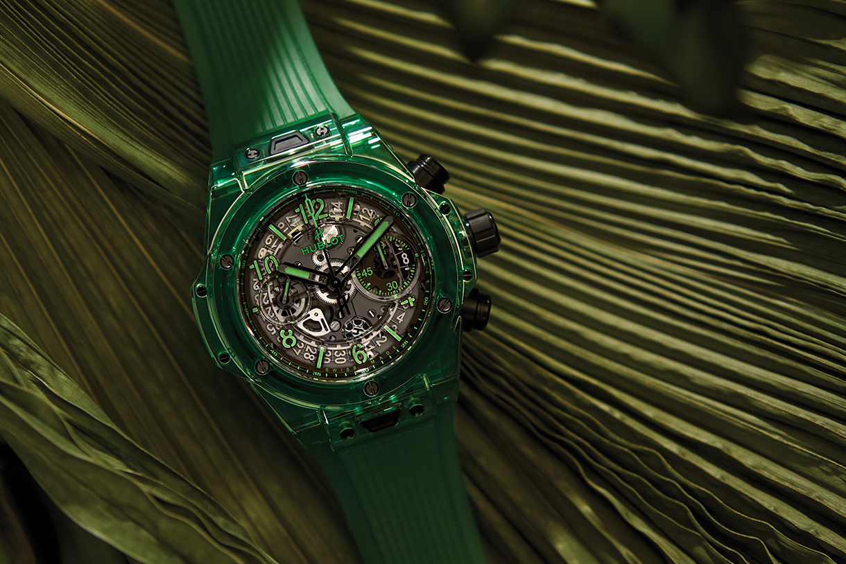 A luxury ceramic chronograph watch with bright green band and black dial resting on a bed of large green leaves.