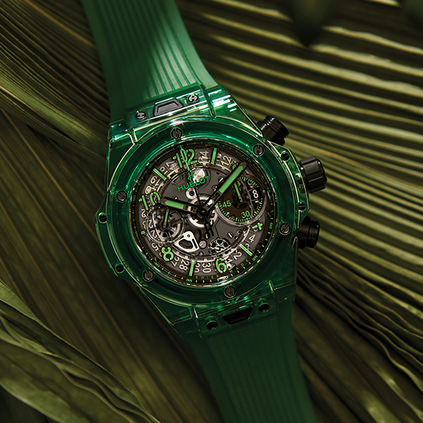 A luxury ceramic chronograph watch with bright green band and black dial resting on a bed of large green leaves.