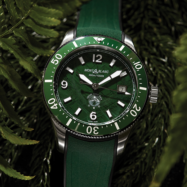 Date watch with green dial, bezel and rubber strap sitting on a pile of green fern leaves.