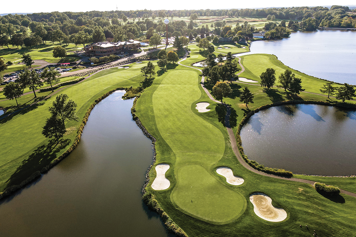  An aerial view of a large, well-manicured golf course. The course is lush green with several lakes and bunkers. A clubhouse and parking lot are visible in the background.