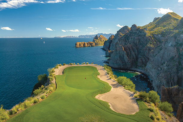 A scenic golf course tee box perched on a cliff overlooking a turquoise ocean. Mountains rise in the background and a sailboat is visible in the distance.