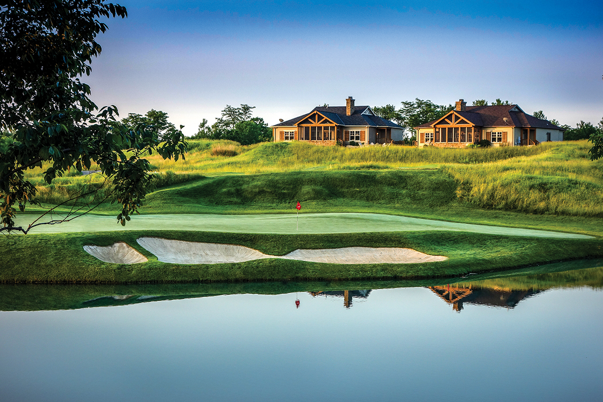  A scenic golf course with a lake and cottages in the background. The green grass is well-manicured, and the water reflects the blue sky and clouds.