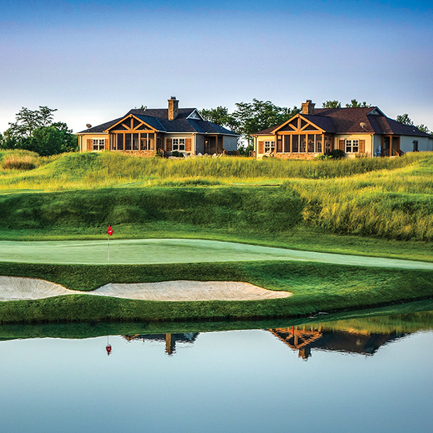 A scenic golf course with a lake and cottages in the background. The green grass is well-manicured, and the water reflects the blue sky and clouds.