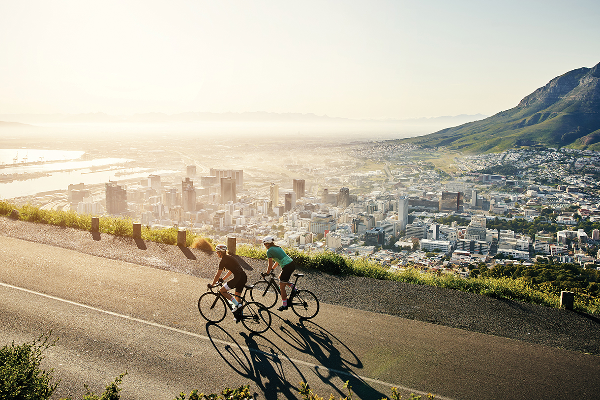 Two people riding bicycles on a paved road overlooking a harbor and city with many tall skyscrapers. There are mountains and ocean in the distance.