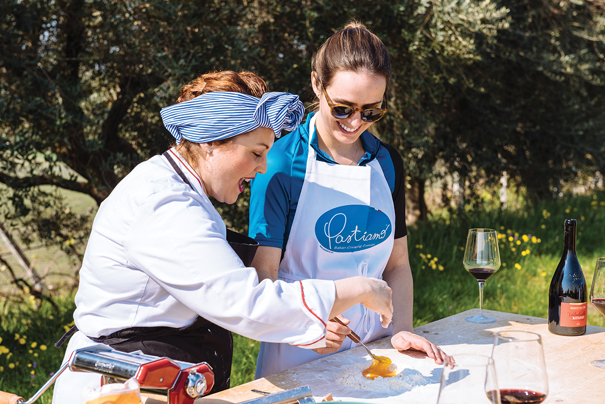 A female chef teaching a woman how to make pasta outdoors. There are glasses of red wine and a pasta machine on the table.