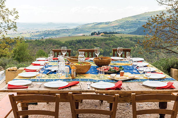 In the foreground, there is a large wooden table set for a meal with glasses, plates, bowls and glasses. In the background there is a view of mountains and houses in Verona, Italy. 