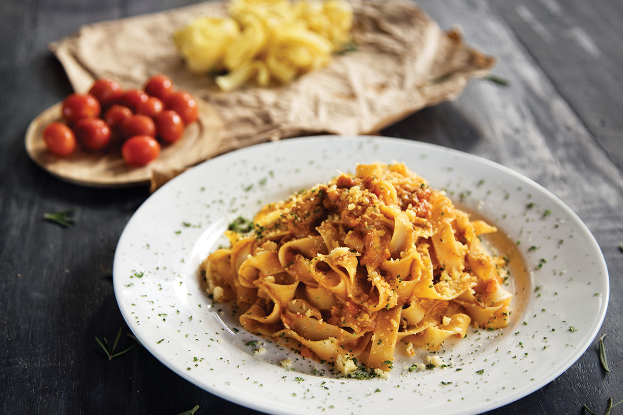 A white plate of tagliatelle pasta with tomato sauce on a wooden table. There is a smaller plate of cherry tomatoes next to it.