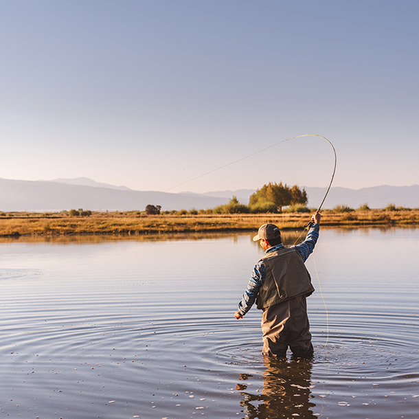 A man is fly fishing in a large, serene pond. He is standing in the middle of the pond, casting a fly rod. The water in the lake is calm and reflects the clear blue sky. There are mountains in the background.