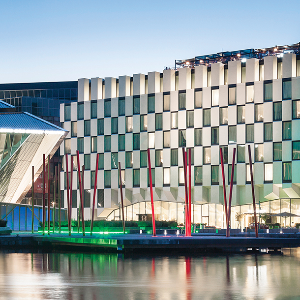 A large, modern glass and steel hotel building with a curved facade, located next to a body of water. There is a dock in front of the building and spotlights lighting up the building from below.