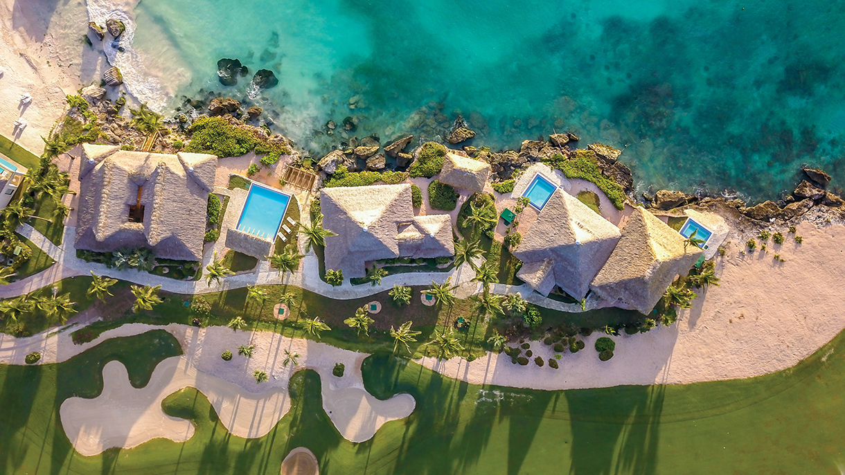 An aerial view of a beach resort with swimming pools, lush green lawns, and palm trees swaying in the breeze. The turquoise water of the ocean stretches out to the horizon.