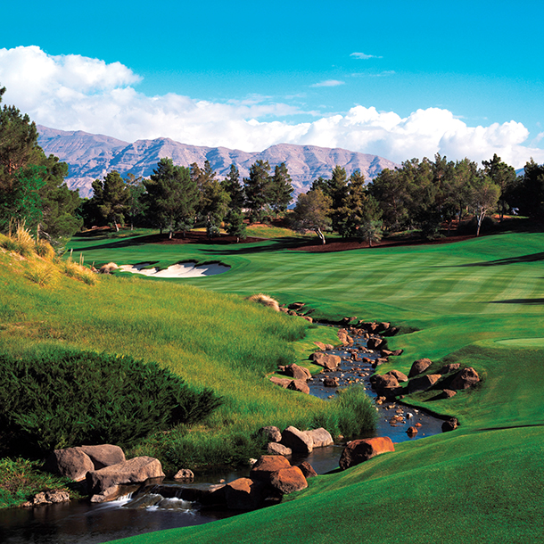A lush green golf course that has a winding fairway and a sand trap on clear day with a very blue sky. There are many trees surrounding the golf course with several mountains visible in the distance.